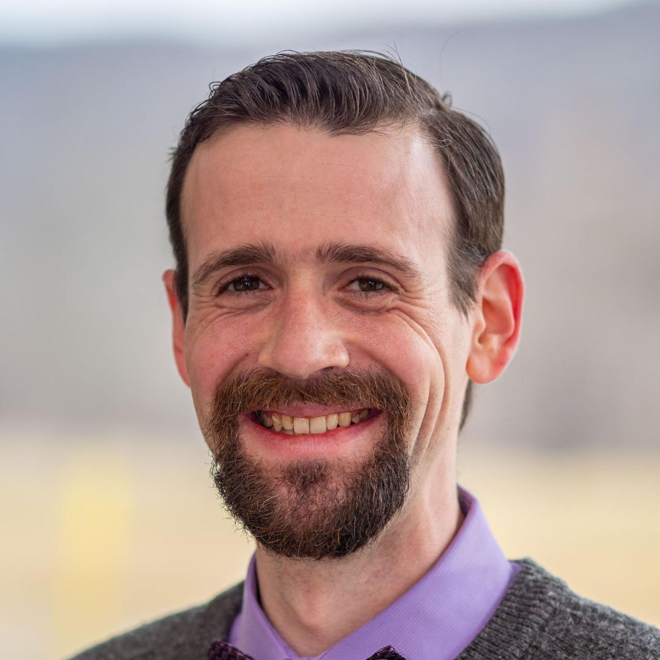 headshot image of a white male with short, combed hair and goatee wearing a purple collared shirt and grey sweater.